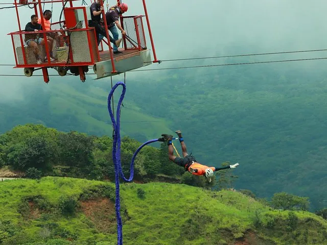 A person bungee jumping from a red suspended tram car over a green valley.